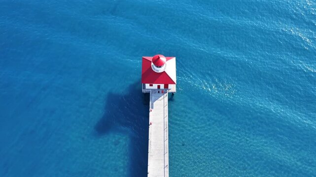 Aerial view of a red roofed lighthouse at the end of a pier casting a giant shadow over the water