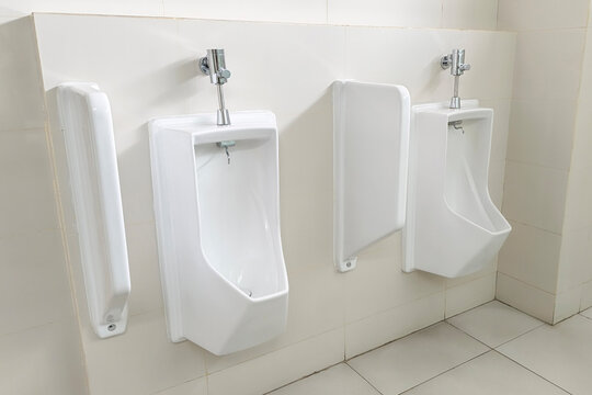 White ceramic urinals in men public restroom interior. Wall mounted male porcelain toilet bowls with flush valve for office building wc, commercial bathroom hygiene, and sanitation concept.