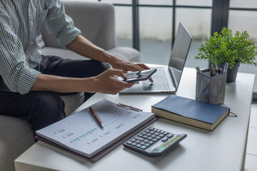 Fototapeta premium Businessman calculating expenses and reviewing financial statistics using smartphone and laptop.