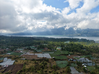 Fototapeta premium Aerial high-angle view of rural farming villages, agricultural fields, and Lake Batur surrounded by green forests under a bright cloudy sky with sunbeams in Bali, Indonesia