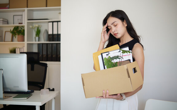 Unemployed woman packing office belongings into a cardboard box after losing her job, looking stressed at her desk.