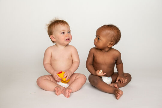 Two babies sitting, one holding a toy, both in diapers on a white background.
