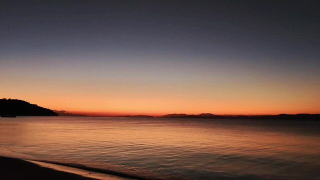 Serene seaside sunset, calm coastal scenest with twilight gradient, transitioning from deep blue at the zenith to vibrant orange and red hues near the horizon. Sea with gentle ripples, golden hour