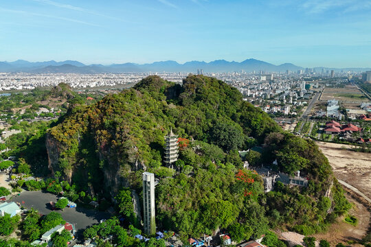 Lush green Marble Mountains, home to pagodas and an elevator, stand tall in Da Nang, Vietnam. A city skyline stretches across the horizon under bright daylight