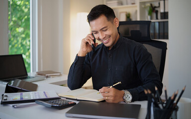 Fototapeta premium Smiling businessman talking on smartphone while writing on financial documents at a bright modern office desk.