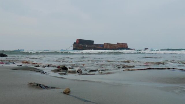 Low-angle shot of ocean waves washing plastic pollution on a beach with a stranded shipwreck background. 