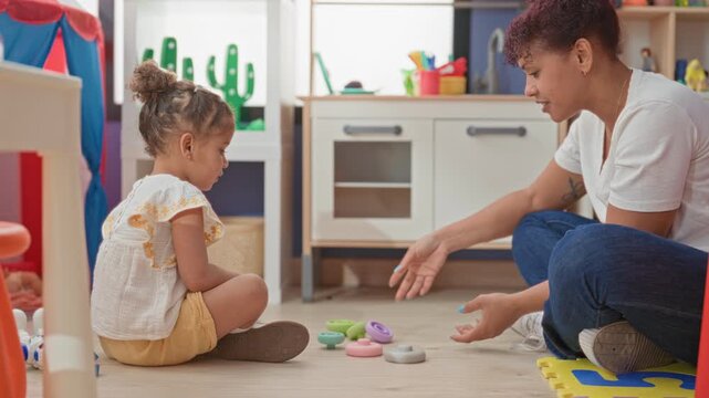 Woman sitting on foam playmat handing stacking ring toy to little girl, hands exchanging pastel rings on classroom floor in building; tender playful learning.