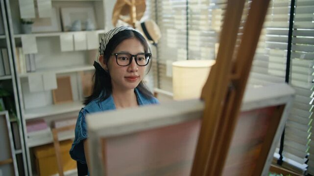 Young Asian woman artist painting on canvas easel with paintbrush in art studio, representing creative professional women designer, fine arts student lifestyle and freelance illustrator