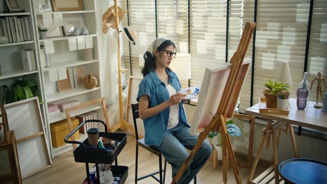 Young Asian woman artist painting on canvas easel with paintbrush in art studio, representing creative professional women designer, fine arts student lifestyle and freelance illustrator