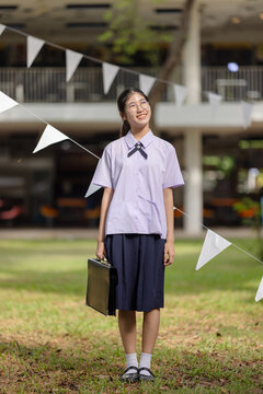 Portrait of a cheerful female student in a traditional Thailand high school uniform, featuring a white short-sleeved shirt with a small bow tie, a navy blue pleated skirt.
