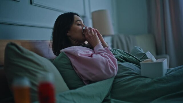 Ill woman taking napkin blowing runny nose in evening bedroom. Unwell lady lying