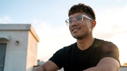 Fototapeta premium Low angle portrait of smiling young man with stylish haircut and clear frame glasses looking away on rooftop at sunset
