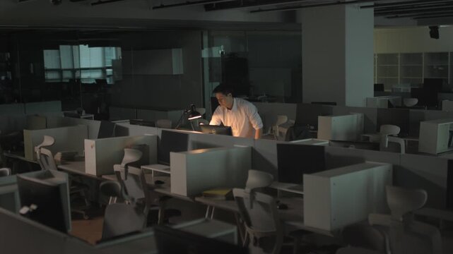 A businessman working late at night alone in a dark office with a single desk lamp illuminating his workstation from the side.