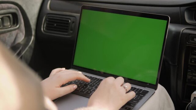 Person types on laptop with green screen inside car. Hands move quickly over keyboard. Focus on typing, not face or emotion. Useful for remote work, travel, or mobile office concepts