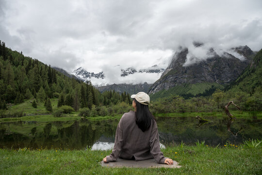 The back view of a woman sitting on the grass by the lake, gazing at the mist-shrouded snow-capped mountains