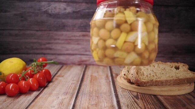 A hand opens a jar of olive next to fresh bread and vegetables