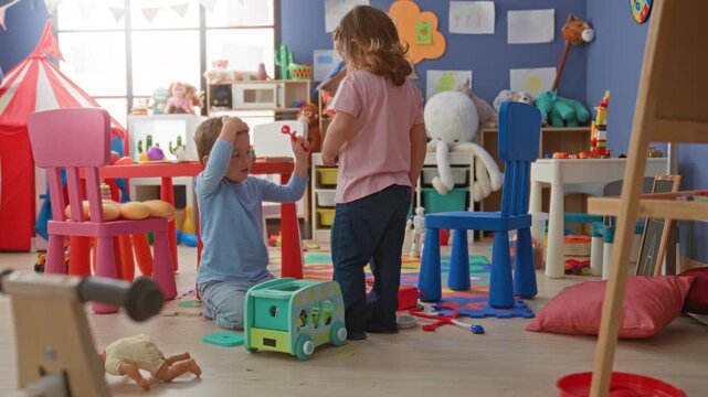 Girl and boy playing with a toy bus, boy seated touching his cheek while girl reaches for blocks amid colorful toys in building playroom; playful curiosity.