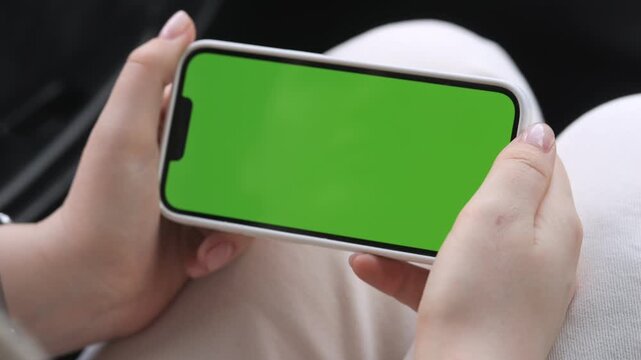 Hands hold smartphone with green screen. Person sits comfortably, focused on device. Ideal for app demos or digital content. Perfect for advertising, tech, or social media use