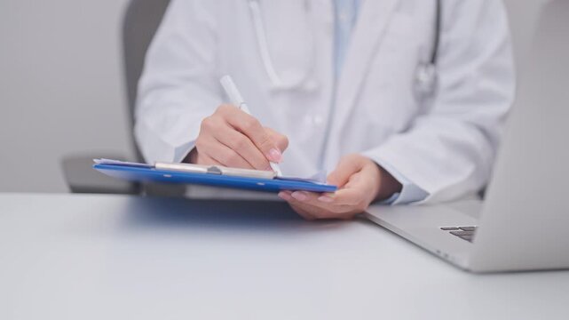 Professional doctor wearing lab coat and stethoscope using laptop computer and taking report notes on notebook at desk in hospital clinic office, medical technology and healthcare concept