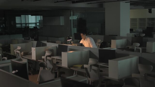 A businessman working late at night at a brightly lit desk in an otherwise completely dark and empty office space.