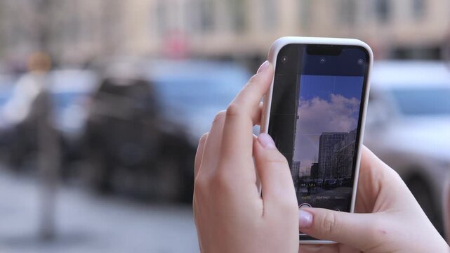 Close-up of hands holding smartphone, capturing urban street scene. Fingers tap screen, adjust camera focus. Background blurred, vehicles and buildings visible