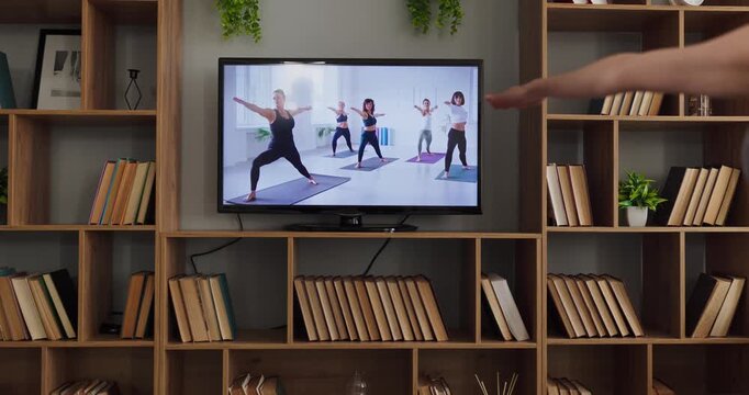 TV yoga class online training fitness home exercise. The TV screen shows a group session as women guide poses. Bookshelves and plants frame calm interior. Concept of guided home fitness.