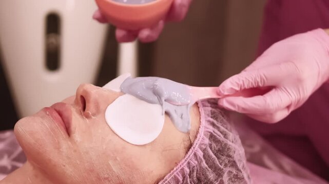 Facial treatment. Detox procedure. Woman in beauty salon with cotton pads on eyes cosmetologist hands applying alginate mask with spatula on face.