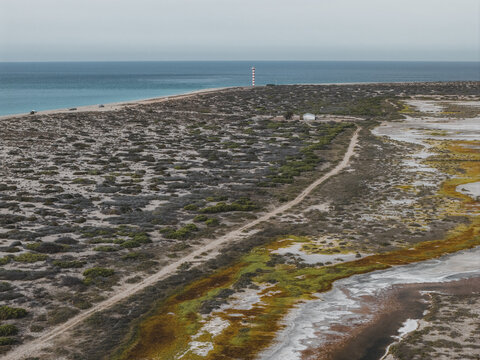 Aerial view of the coastline featuring a red and white striped lighthouse, desert vegetation, a dirt path, and tidal flats along the sea in La Ventana, Baja California Sur, Mexico.