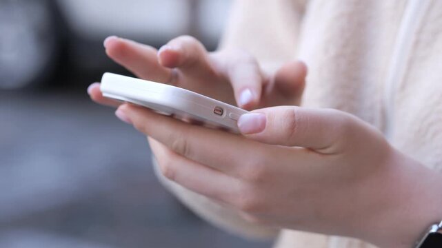 Close-up of hands holding a white smartphone. Fingers gently tap the screen. Person wears light-colored sweater. Background softly blurred. Suitable for tech, communication, or daily life themes