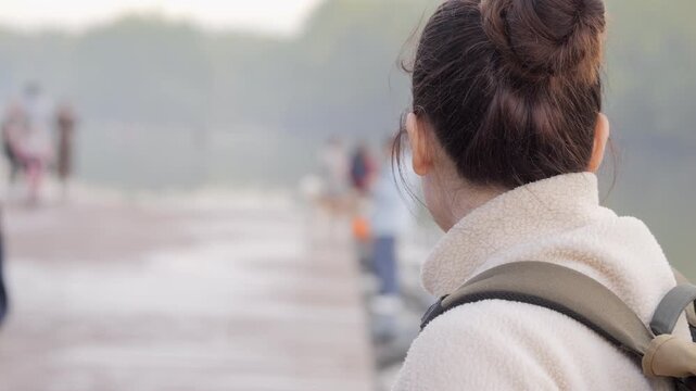Woman walks on bridge, back view, hair in bun, wearing cozy jacket and backpack. Background blurred with people and nature. Calm, reflective mood, suitable for travel, wellness, or meditation content