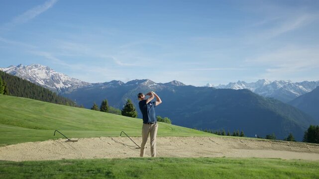 Skilled golfer blasts a shot from a sand bunker on a mountain golf course in Verbier, Switzerland. Slow motion sand spray with dramatic alpine peaks in the background.