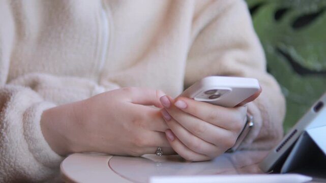 Close-up of hands typing on a white smartphone. Person wears soft beige sweater. Fingers move quickly over screen, showing focus. Background has green leafy plant