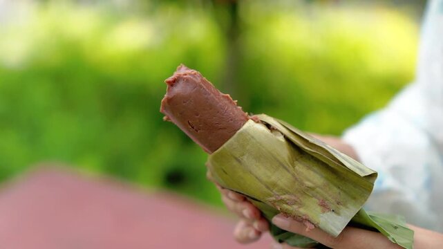 A young Asian Muslim woman is enjoying getuk pisang, a specialty food from Kediri, Indonesia, made from mashed steamed bananas wrapped in banana leaves.