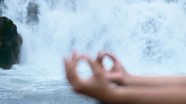 Close-up of hands performing meditation mudra with blurred natural waterfall background for relaxation and mindfulness practice outdoors in calm environment