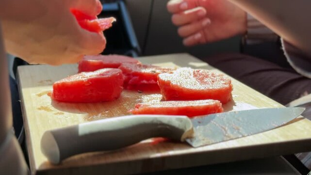 Close-up of hands using a knife to slice fresh red watermelon on a wooden cutting board inside a car. Bright sunlight illuminates the juicy fruit during a summer road trip or picnic stop.