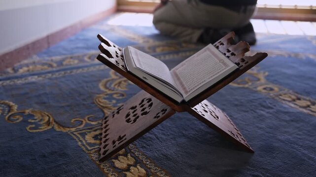 An open Holy Quran rests on a carved wooden stand indoors. In the blurred background, a person kneels in prayer on a blue patterned carpet inside a softly lit mosque during bright daylight.