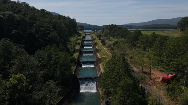 Aerial View Cascade Spillway At Da Lat Vietnam, Pine Forest Flanking Concrete Channel, Stepped Pools Pouring Clear Blue Water Toward Mountain Lake, Sunlight Dappling Treetops, Calm Summer