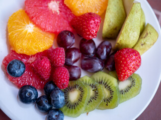 Obraz premium Top view of fresh mixed fruits on a white plate, including kiwi, strawberries, blueberries, raspberries, grapes, pear, orange and grapefruit slices.