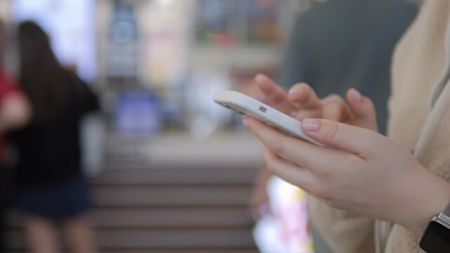 Close-up of hands holding and using a smartphone. Fingers tap screen while walking in public space. Blurred background shows people moving. Ideal for mobile usage, urban lifestyle