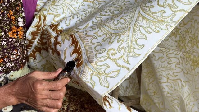 Close-up video of artisan hands drawing intricate golden floral batik patterns on white fabric using a canting tool, representing traditional Indonesian batik craft