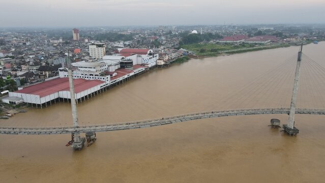 Aerial view of Jembatan Gentala Arasy spanning the Batang Hari River with urban buildings and rooftops along the riverbank in Kecamatan Jambi Timur, Jambi, Indonesia.