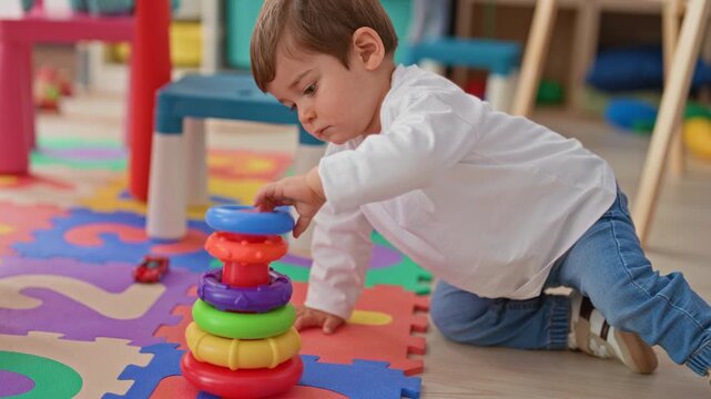 Boy stacking colorful ring toy on foam playmat, hand pressing top ring while kneeling in building; curiosity learning.