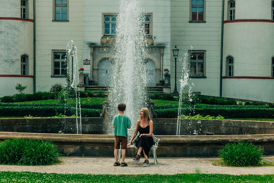 Mother with little son sitting on bench by fountain in park near castle, Europe, summer.