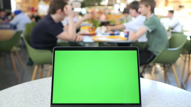 Laptop with green screen sits on cafe table. Background shows blurred people chatting and eating. Ideal for mockups or presentations needing focus on screen content