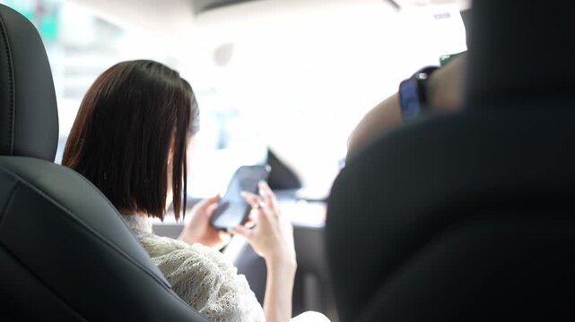 Young woman sits, focused on her phone inside a car. She holds the device, eyes fixed on its screen. Soft light filters through the car windows. Passenger beside her rests their arm on the seat