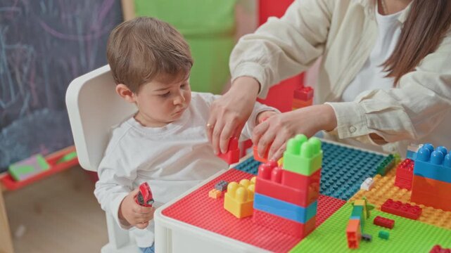 Woman guides little boy stacking colorful toy blocks with hands on tabletop in building; early learning playful development.