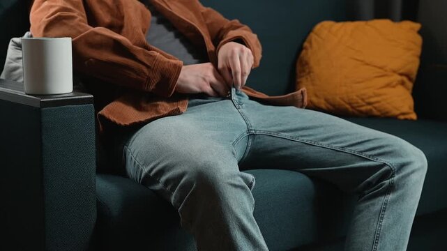 An unrecognizable adult man unzips his fly and puts his hand down his pants while sitting on a sofa in a room