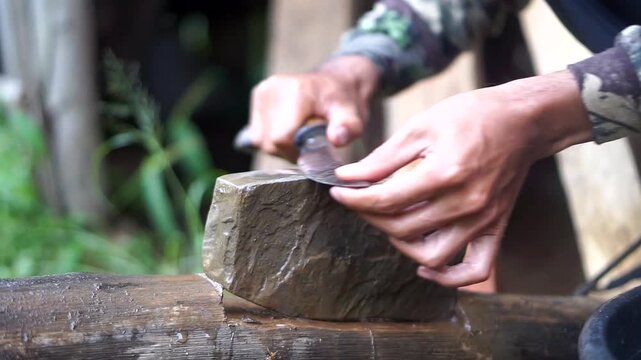 Close-up shot of a man's hands sharpening a traditional kitchen knife blade using a wet natural whetstone on a wooden block.