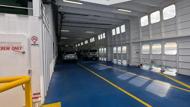 Parked cars line the blue vehicle deck inside a moving passenger ferry with sea and sky visible through side windows.