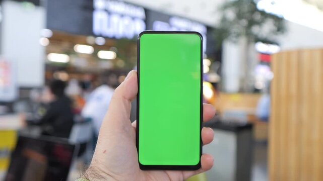 Hand holds phone with green screen. Blurred background shows indoor public space. Focus on device for app or design mockups. Ideal for digital interfaces, ads, or UI concepts